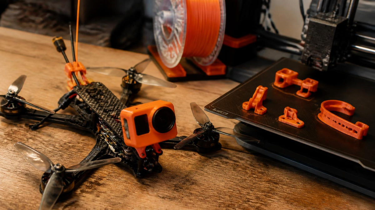 FPV drone with an orange 3D printed TPU camera mount next to a 3D printer bed of freshly printed orange parts