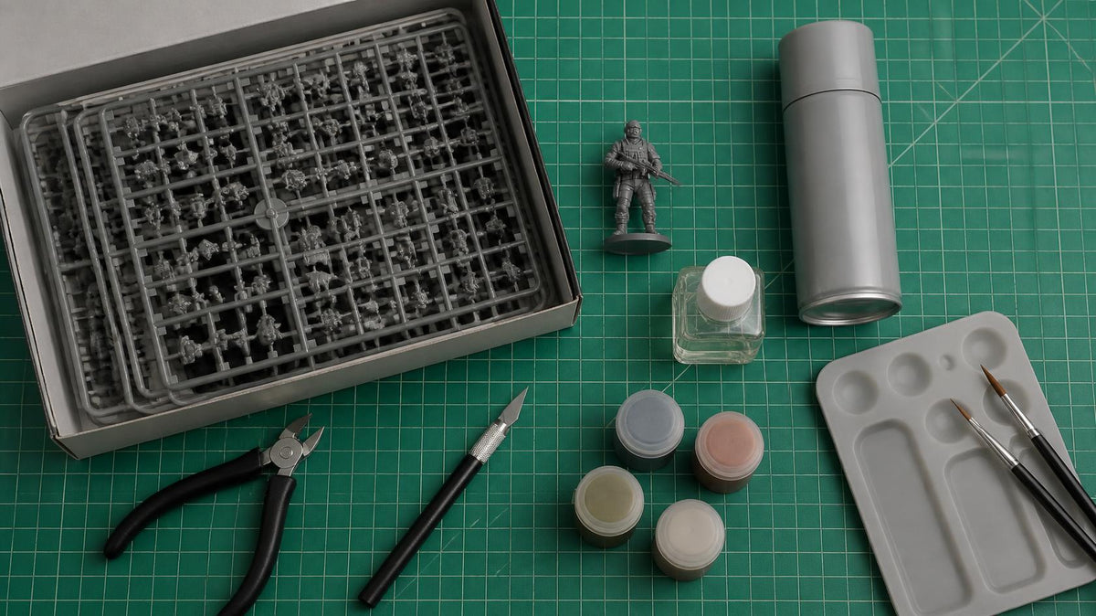 Box of miniature sprues with clippers, hobby knife, plastic cement, spray primer, paint pots, brushes and a palette on a cutting mat